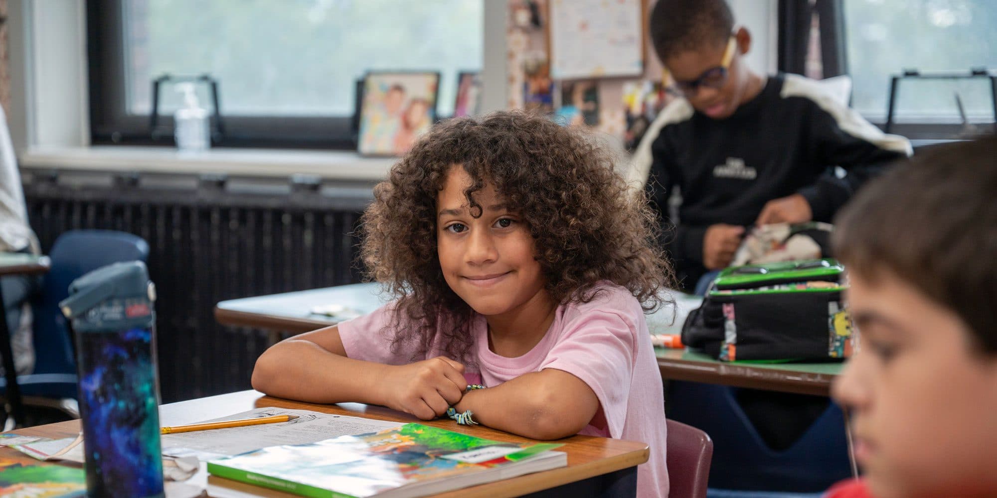 Smiling student with curly hair seated at a classroom desk with books.