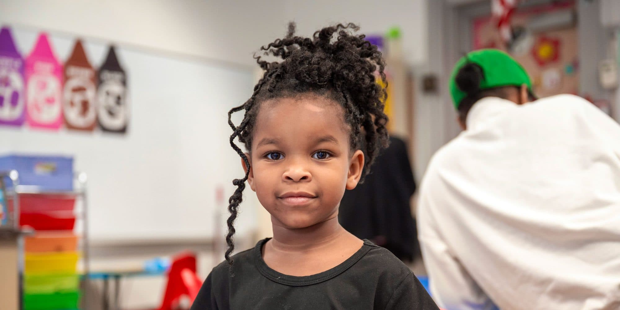 Young student with curly hair smiling in a colorful classroom.