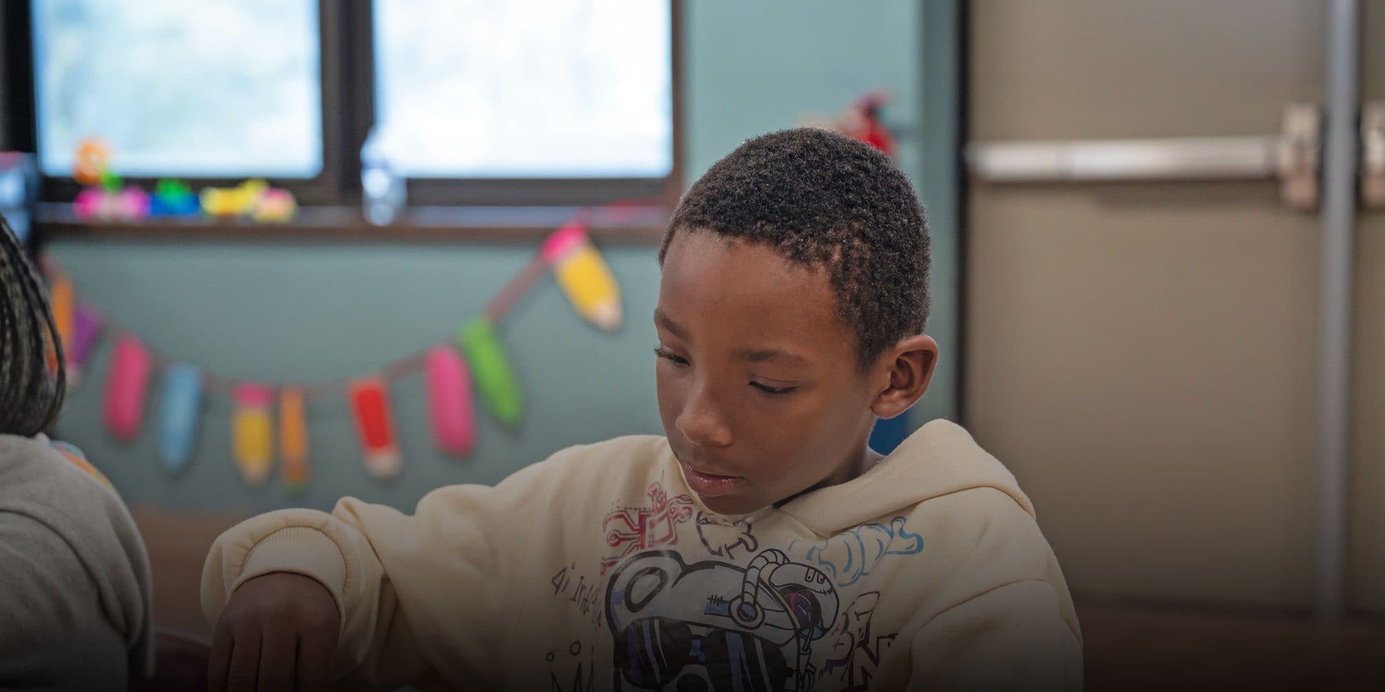Student in a light hoodie focused on work at a classroom desk.