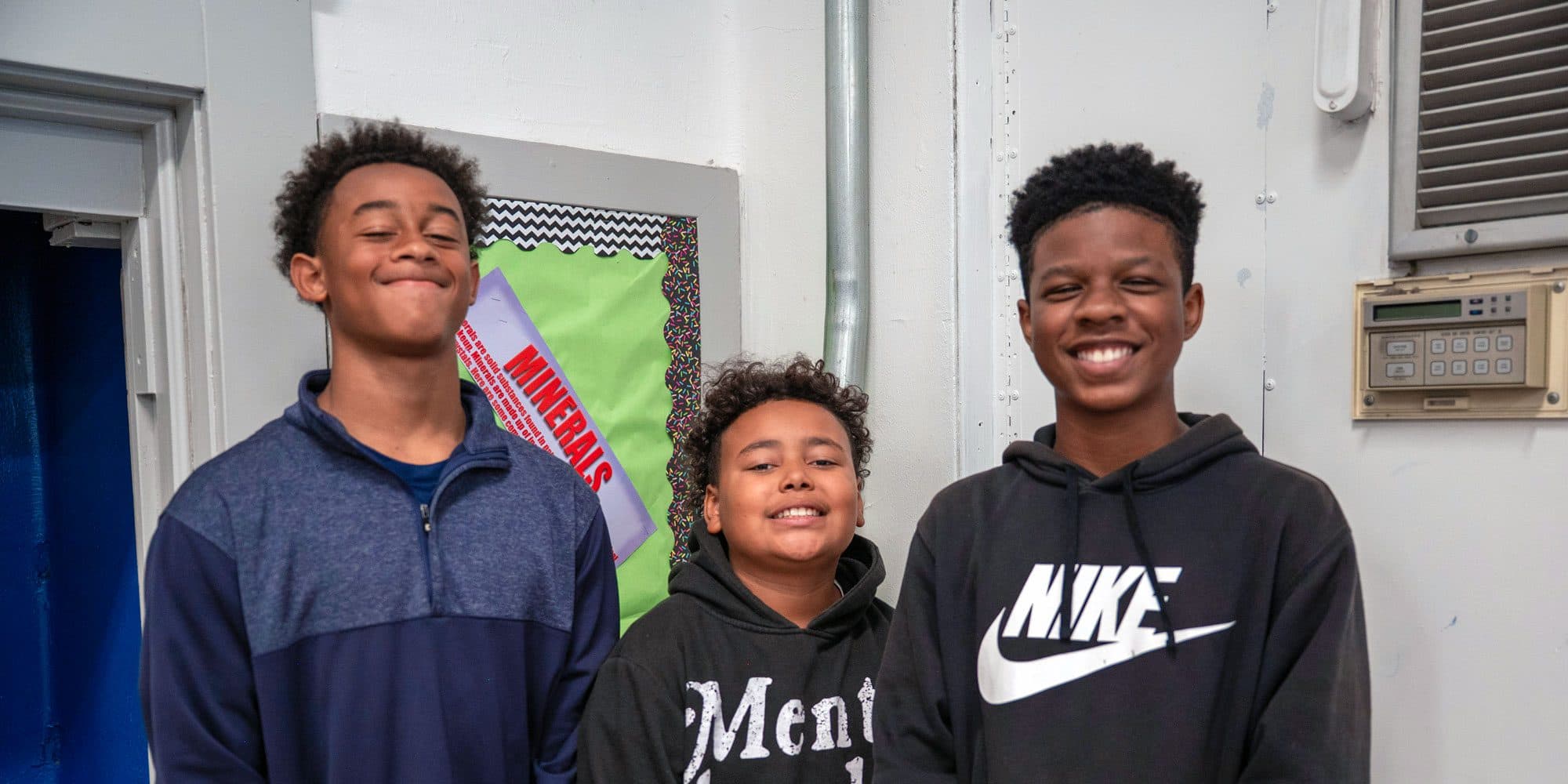 Three smiling students standing together in front of a classroom bulletin board.